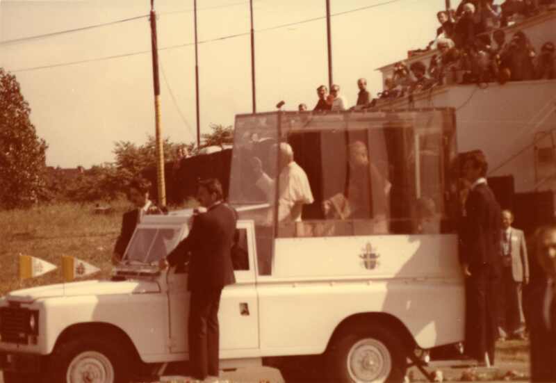 Jan Paweł II w otoczeniu ochroniarzy wjeżdża papamobile na teren Parku Kultury (Łęgi Dębińskie, obecnie Park Jana Pawła II). Na drugim planie wierni na tarasie domu. 20 VI 1983 r. Sygnatura: IPNPo-7-7-2-15